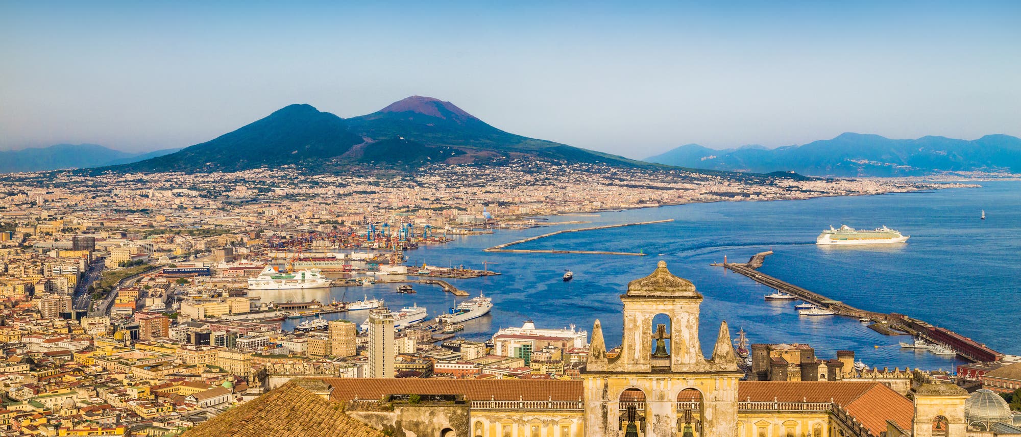 Stadtansicht von Neapel mit dem Vesuv im Hintergrund. Im Vordergrund sind historische Gebäude mit Glockenturm zu sehen. Der Hafen ist mit Kreuzfahrtschiffen und Frachtschiffen belebt. Der Himmel ist klar und blau, was auf einen sonnigen Tag hinweist.