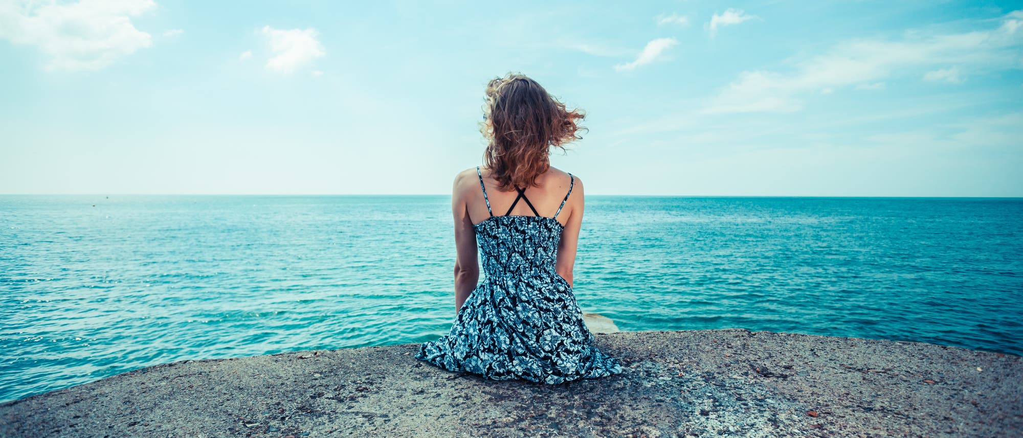 Eine Frau im blauen Kleid, von hinten fotografiert, wie sie aufs Meer hinaus schaut.