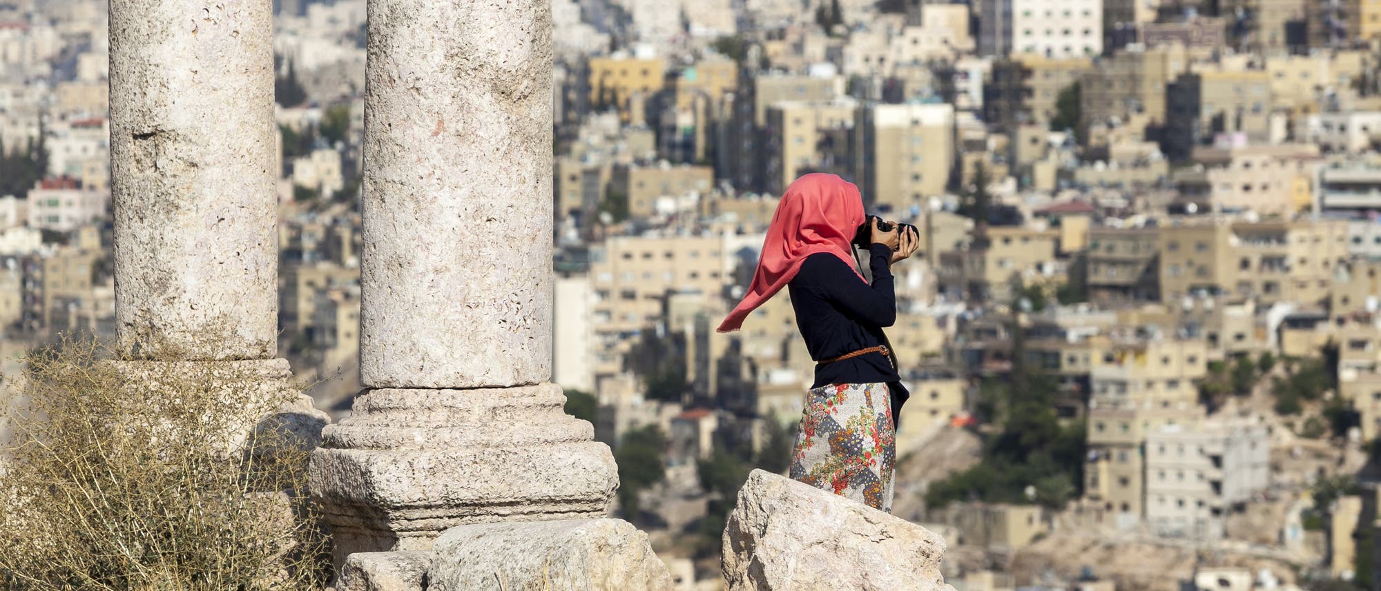 Eine junge Frau mit rotem Kopftuch steht mit einer Kamera filmend neben zwei römischen Säulenstümpfen vor den Häusern von Amman