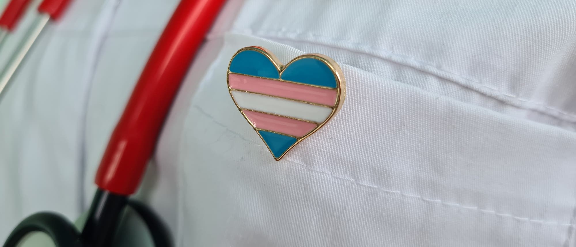 Ein Stethoskop mit roten Schläuchen hängt um den Hals einer Person in einem weißen Kittel. An der Brusttasche des Kittels ist ein herzförmiger Anstecker mit den Farben Blau, Rosa und Weiß befestigt, der die Transgender-Flagge darstellt.