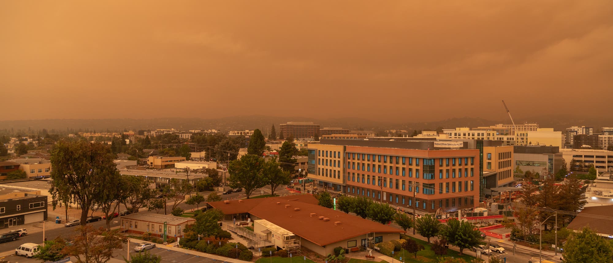 Städtische Landschaft unter einem orangefarbenen Himmel, der durch Rauch von Waldbränden verursacht wird. Im Vordergrund sind Gebäude und ein Parkplatz zu sehen, während im Hintergrund weitere städtische Strukturen und Bäume sichtbar sind. Die Atmosphäre wirkt düster und ungewöhnlich gefärbt. Städtische Landschaft unter einem orangefarbenen Himmel, der durch Rauch von Waldbränden verursacht wird. Im Vordergrund sind Gebäude und ein Parkplatz zu sehen, während im Hintergrund weitere städtische Strukturen und Bäume sichtbar sind. Die Atmosphäre wirkt düster und ungewöhnlich gefärbt.