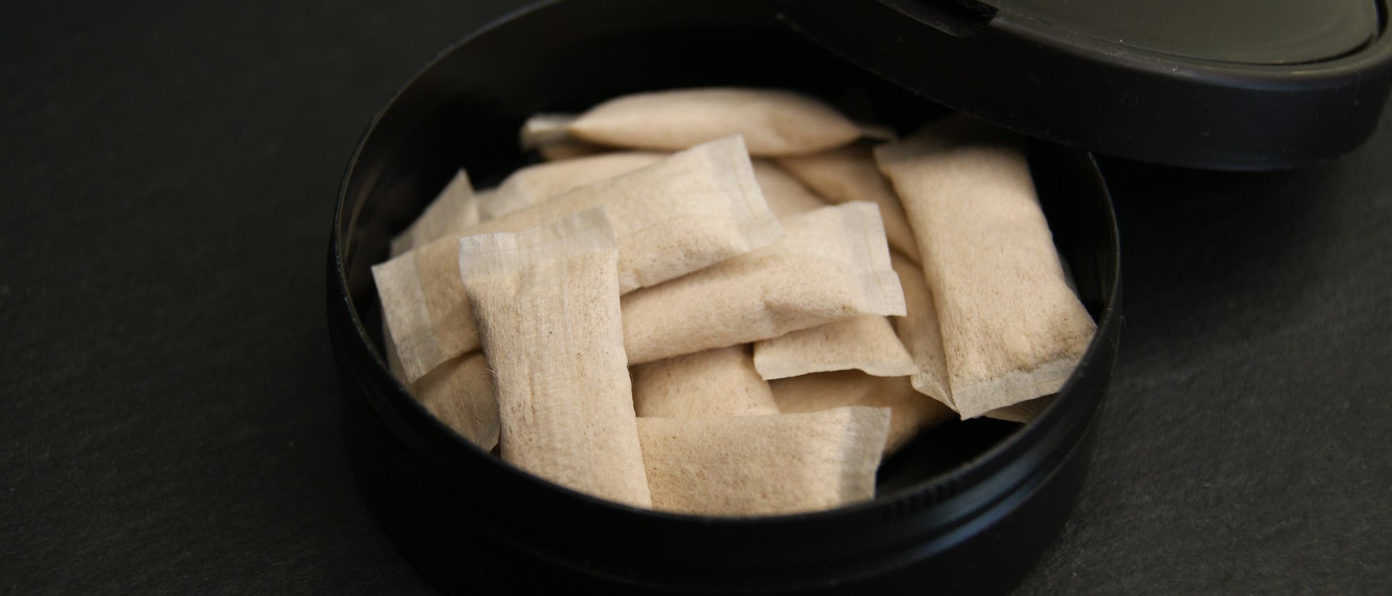 Eine offene schwarze Dose mit mehreren weißen Beuteln, die Nikotin enthalten, auf einer dunklen Oberfläche. Die Beutel sind ordentlich gestapelt und die Dose ist teilweise geöffnet. Eine offene schwarze Dose mit mehreren weißen Beuteln, die Nikotin enthalten, auf einer dunklen Oberfläche. Die Beutel sind ordentlich gestapelt und die Dose ist teilweise geöffnet.