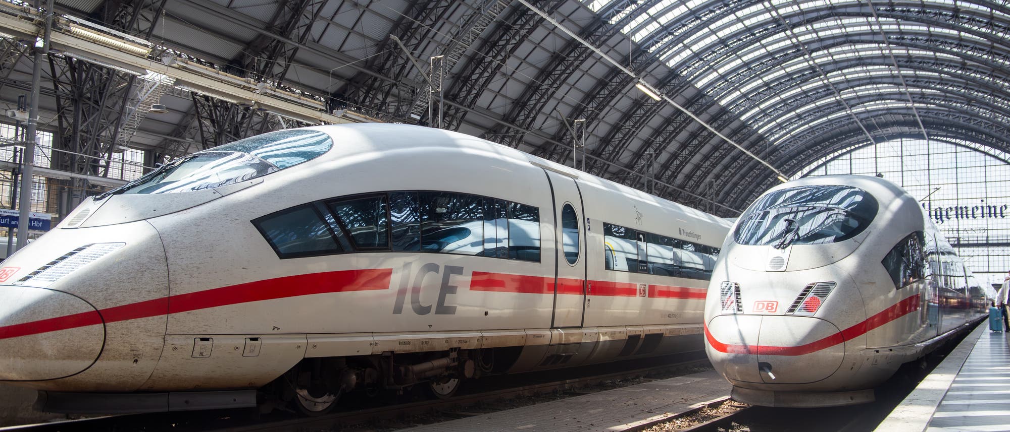 Zwei Hochgeschwindigkeitsz&uuml;ge stehen nebeneinander in einem &uuml;berdachten Bahnhof. Die Z&uuml;ge sind wei&szlig; mit roten Streifen und tragen das Logo der Deutschen Bahn. Die Bahnhofshalle hat eine gew&ouml;lbte Glas- und Stahlkonstruktion, durch die Sonnenlicht einf&auml;llt. Im Hintergrund sind Gleise und Bahnsteige zu sehen.