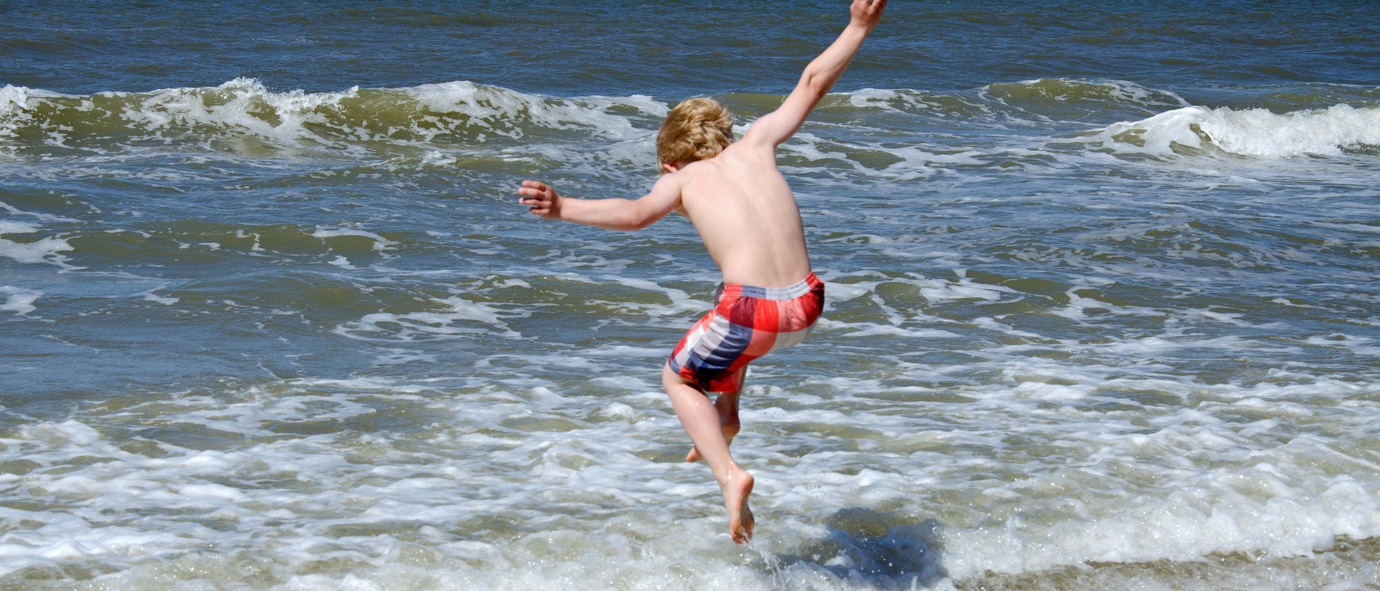 Man sieht ein Kind am Strand von hinten in einer rot-blau gestreiften Badehose, das freudig in die Wellen springt. Der Himmel ist klar und blau, das Meer zeigt leichte Wellen. Die Szene vermittelt ein Gefühl von Sommer und Unbeschwertheit.