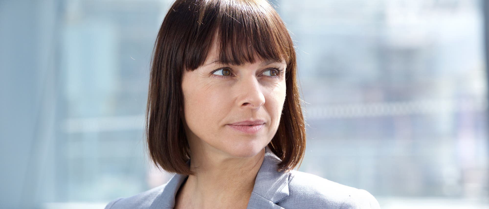 Business Frau im Blazer vor typischer (Stahl-Glas-)Bürogebäudefassade in Unschärfe im Hintergrund.