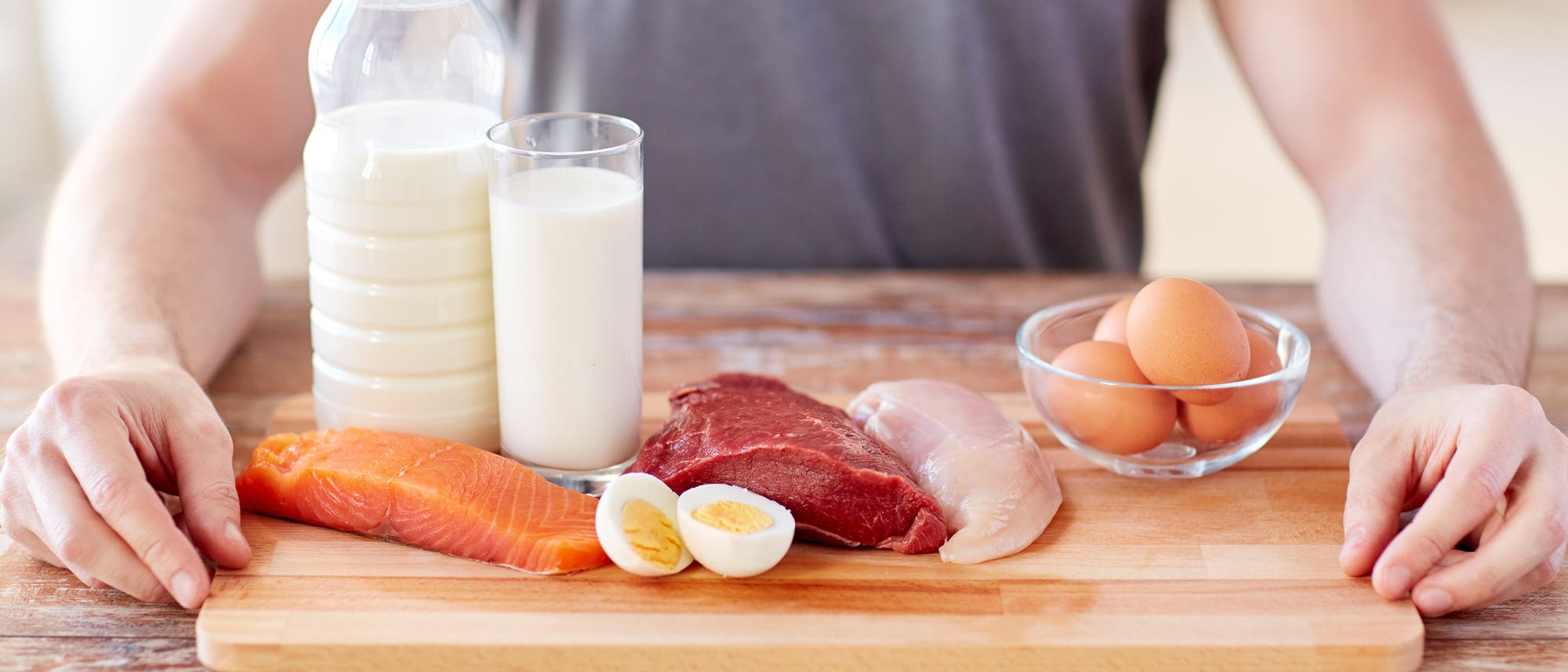 Ein Brett mit allerlei Lebensmitteln, die Proteine enthalten (Milch, Käse, Wurst, Eier, Fisch), im Hintergrund der Oberkörper eines Menschen, die Arme rechts und links des Tabletts.