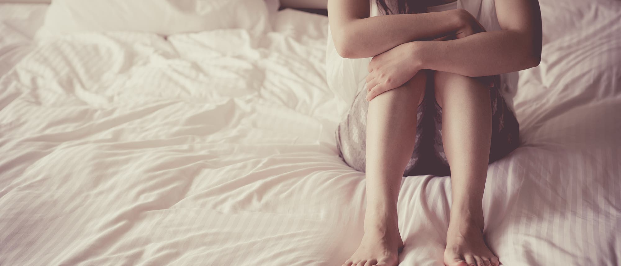 Eine Frau sitzt in kurzer Jeans und Hemd auf dem Bett, das weiß bezogen ist.