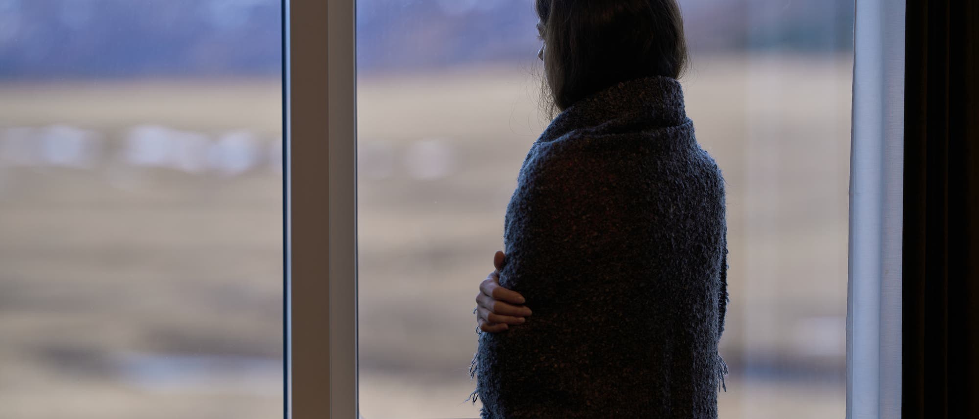 Eine Person steht in einem Raum und blickt aus einem großen Fenster auf eine verschwommene Landschaft. Sie ist in eine dunkle Decke gehüllt und scheint nachdenklich. Draußen sind Felder und Berge zu erkennen. Eine Person steht in einem Raum und blickt aus einem großen Fenster auf eine verschwommene Landschaft. Sie ist in eine dunkle Decke gehüllt und scheint nachdenklich. Draußen sind Felder und Berge zu erkennen.