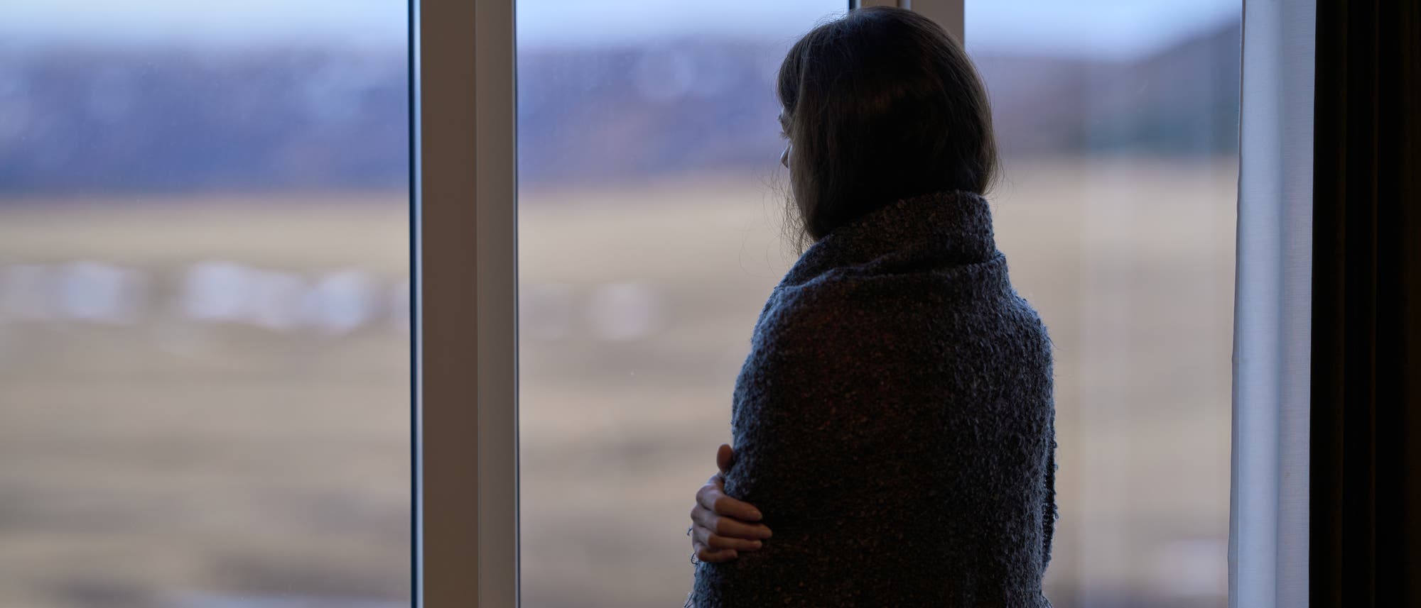 Eine Person steht in einem Raum und blickt aus einem großen Fenster auf eine verschwommene Landschaft. Sie ist in eine dunkle Decke gehüllt und scheint nachdenklich. Draußen sind Felder und Berge zu erkennen. Eine Person steht in einem Raum und blickt aus einem großen Fenster auf eine verschwommene Landschaft. Sie ist in eine dunkle Decke gehüllt und scheint nachdenklich. Draußen sind Felder und Berge zu erkennen.