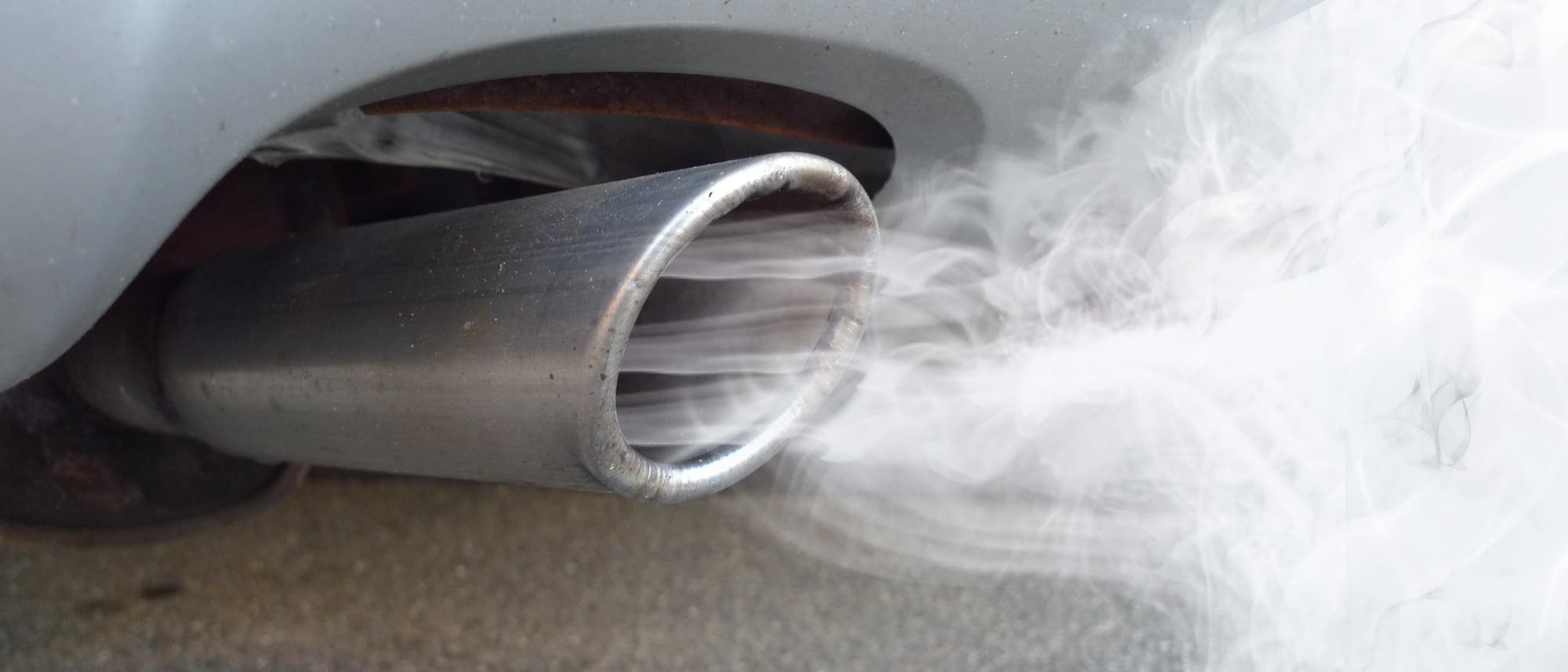Ein Autoauspuff, aus dem weißer Rauch austritt, vor einem grauen Hintergrund. Der Rauch deutet auf die Emissionen des Fahrzeugs hin.