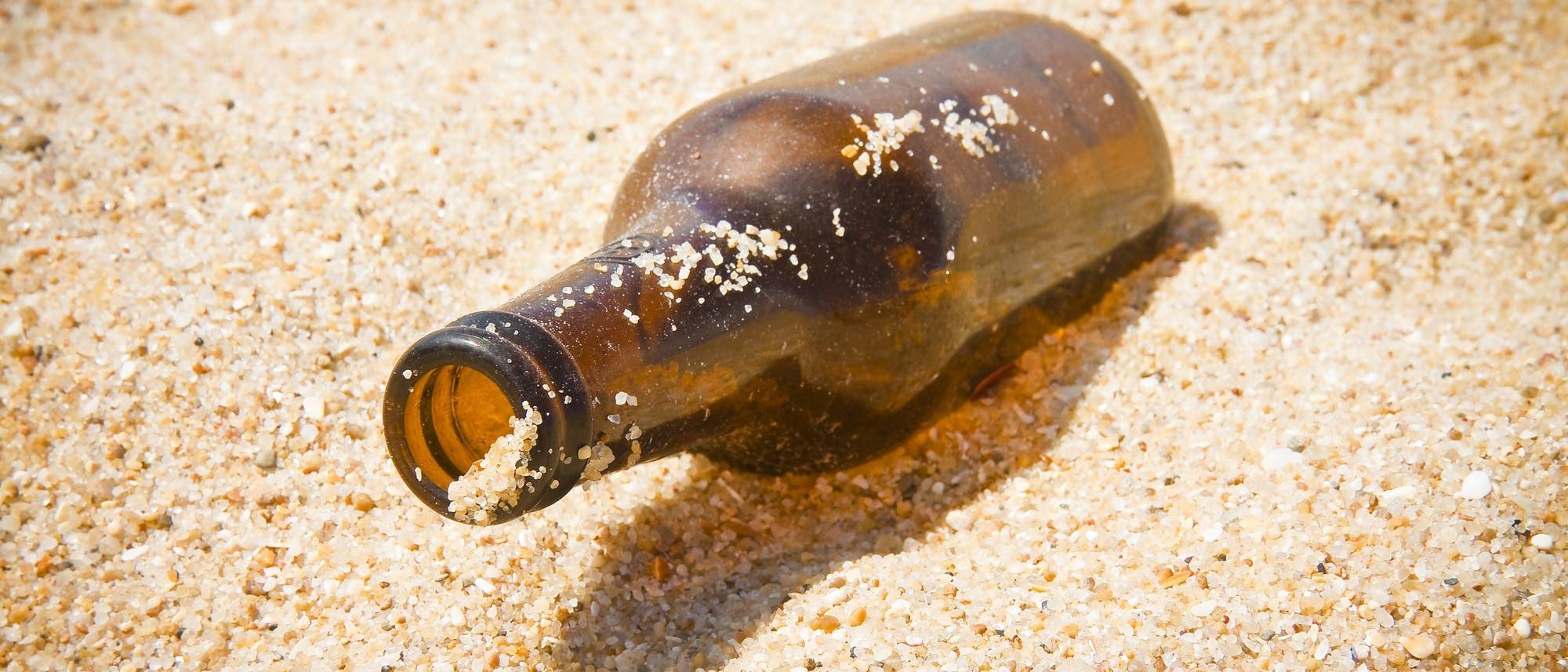 Eine leere braune Flasche ohne Etikett liegt im Sand.