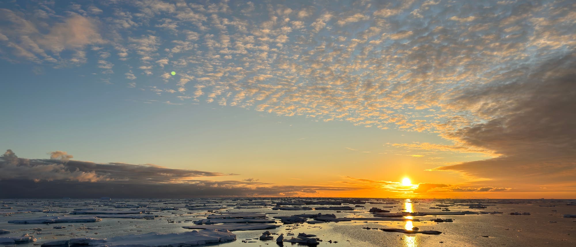 Sonnenaufgang über lückenhaftem Meereis in der Antarktis. Die Sonne leuchtet rotorange, der Himmel zeigt verschiedene Schattierungen von hell- bis dunkelblau. Das Meer wirkt düster, am Himmel sind einzelne Wolken