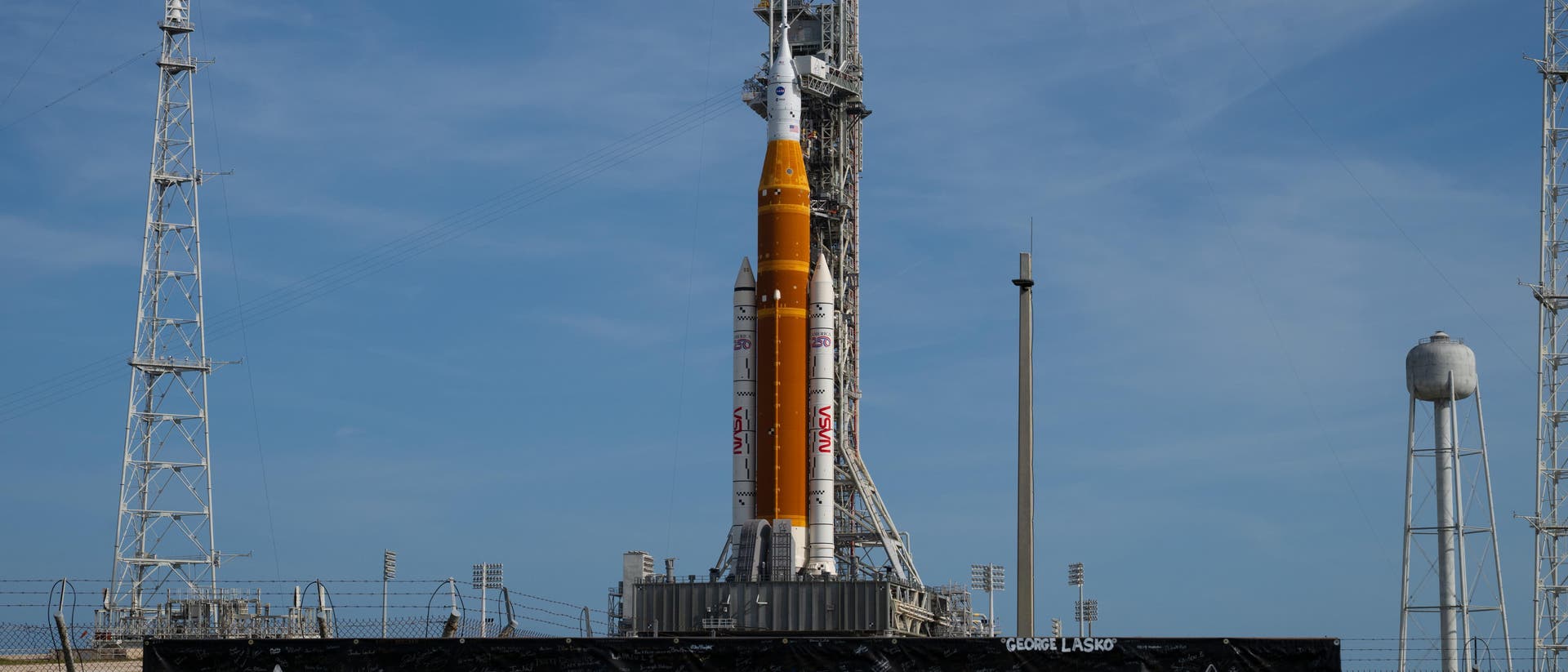 Eine gro&szlig;e Rakete steht auf einer Startrampe, umgeben von T&uuml;rmen und einem Wasserturm. Im Vordergrund ist ein Banner mit dem Text "LET'S GO!" und dem Logo der Artemis-Mission zu sehen. Der Himmel ist klar und blau.