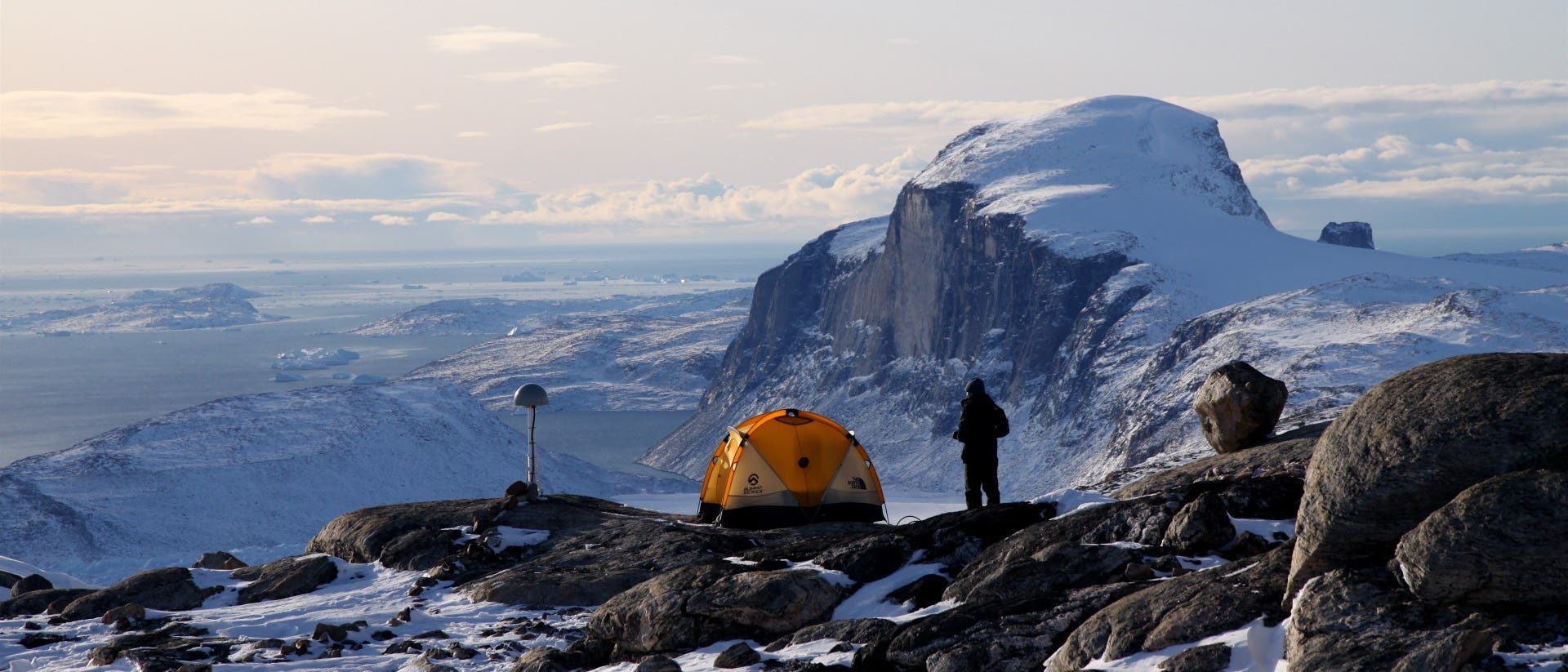 Ein in Schwarz gekleideter Forscher steht neben einem gelben Kuppelzelt und einer Messstation auf einem Berg nahe der Küste Grönlands. Die Landschaft ist mit Schnee und Eis bedeckt