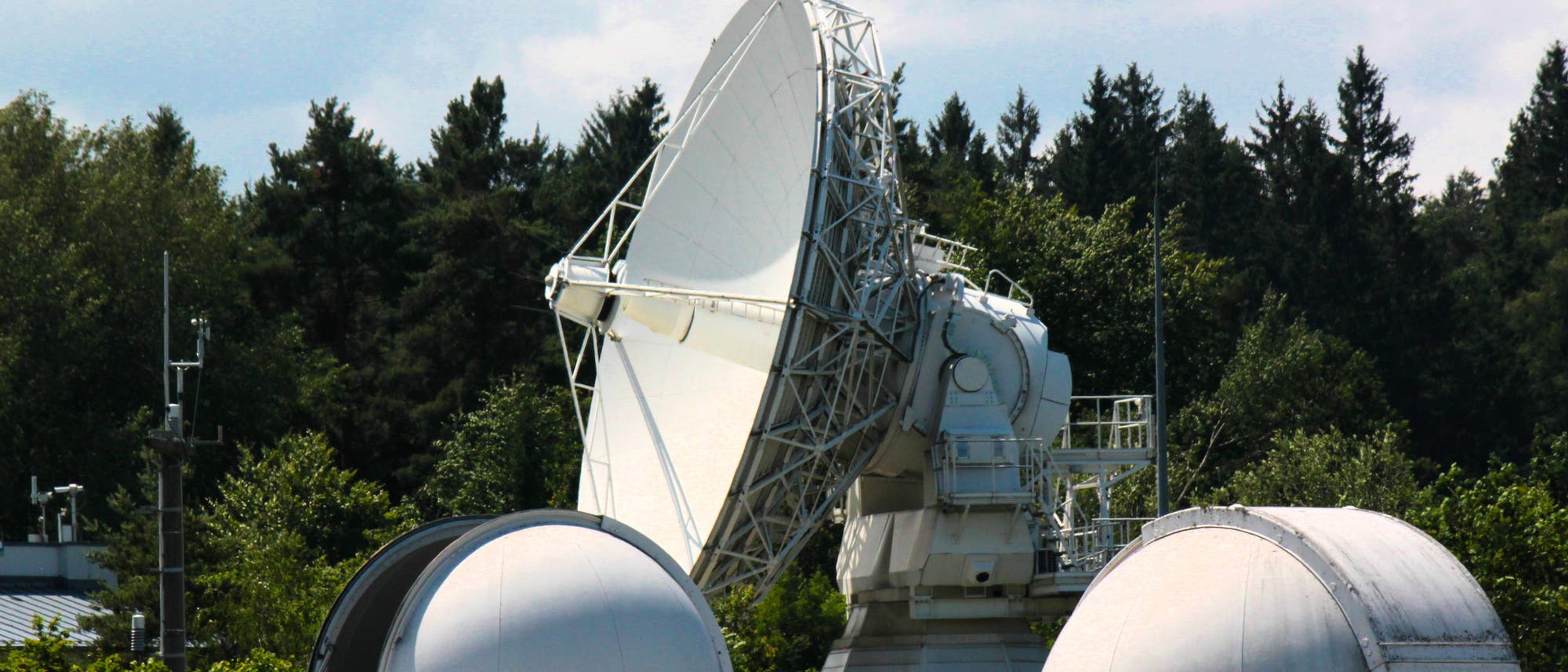 Eine große Satellitenschüssel und zwei kleinere Kuppeln eines Observatoriums sind von Bäumen umgeben. Die Satellitenschüssel ist auf einem stabilen Sockel montiert und zeigt in den Himmel. Die Kuppeln sind teilweise geöffnet, was auf eine aktive Nutzung hinweist. Im Hintergrund sind dichte Wälder und ein bewölkter Himmel zu sehen. Eine große Satellitenschüssel und zwei kleinere Kuppeln eines Observatoriums sind von Bäumen umgeben. Die Satellitenschüssel ist auf einem stabilen Sockel montiert und zeigt in den Himmel. Die Kuppeln sind teilweise geöffnet, was auf eine aktive Nutzung hinweist. Im Hintergrund sind dichte Wälder und ein bewölkter Himmel zu sehen.