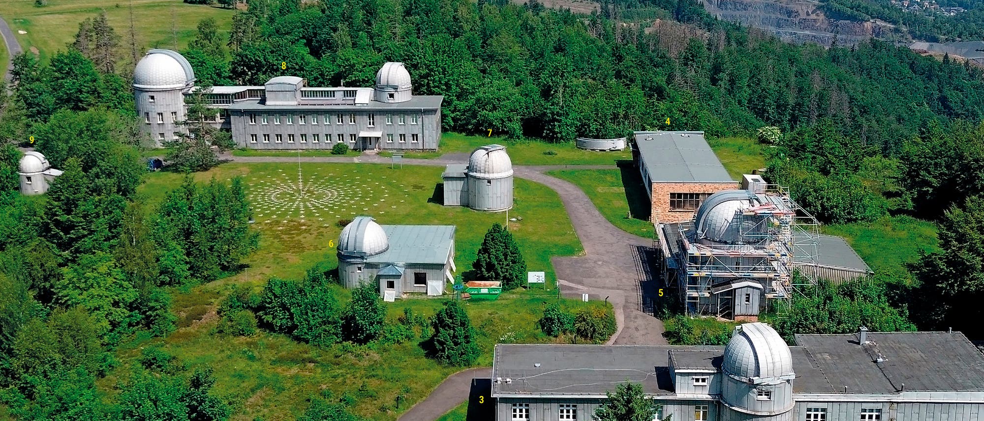 Die Luftaufnahme zeigt die Sternwarte Sonneberg eingebettet in eine grüne, hügelige Landschaft. Mehrere Gebäude mit Kuppeln, die Teleskope beherbergen, sind auf dem Gelände verteilt. Ein großes Hauptgebäude steht zentral, umgeben von kleineren Gebäuden und Kuppeln. Die Gebäude tragen Nummern. Die Umgebung ist dicht bewaldet. Wege verbinden die verschiedenen Teile des Observatoriums. Die Luftaufnahme zeigt die Sternwarte Sonneberg eingebettet in eine grüne, hügelige Landschaft. Mehrere Gebäude mit Kuppeln, die Teleskope beherbergen, sind auf dem Gelände verteilt. Ein großes Hauptgebäude steht zentral, umgeben von kleineren Gebäuden und Kuppeln. Die Gebäude tragen Nummern. Die Umgebung ist dicht bewaldet. Wege verbinden die verschiedenen Teile des Observatoriums.
