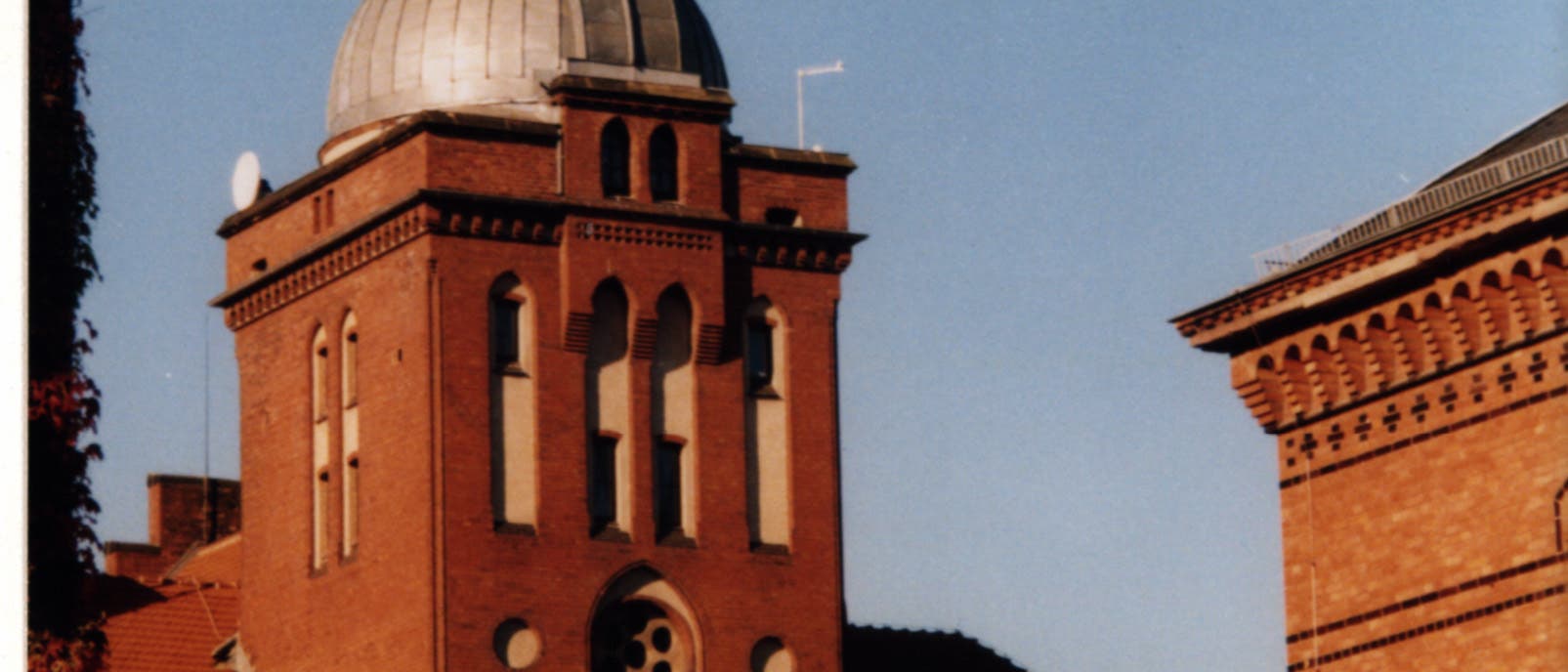 Ein historisches Gebäude aus rotem Backstein mit einem markanten Turm und einer Kuppel, die an ein Observatorium erinnert. Der Himmel ist klar und blau, und das Gebäude ist von weiteren architektonischen Strukturen umgeben. Im Vordergrund sind Bäume zu sehen, die teilweise das Gebäude verdecken. Ein historisches Gebäude aus rotem Backstein mit einem markanten Turm und einer Kuppel, die an ein Observatorium erinnert. Der Himmel ist klar und blau, und das Gebäude ist von weiteren architektonischen Strukturen umgeben. Im Vordergrund sind Bäume zu sehen, die teilweise das Gebäude verdecken.