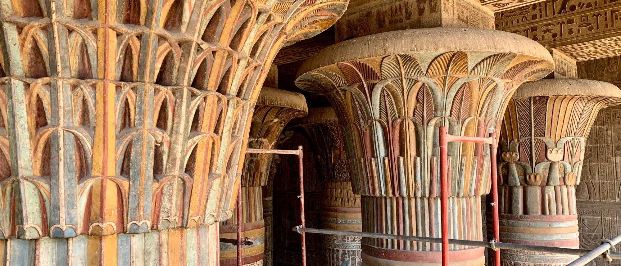 Mehrere reich verzierte, farbenfrohe Säulen im Tempel von Hathor in Dendera, Ägypten. Die Säulen sind mit floralen Mustern und Hieroglyphen verziert. Im Hintergrund sind weitere Säulen und Wände mit antiken ägyptischen Inschriften zu sehen. Ein Gerüst ist zwischen den Säulen aufgebaut, was auf Restaurierungsarbeiten hindeutet. Mehrere reich verzierte, farbenfrohe Säulen im Tempel von Hathor in Dendera, Ägypten. Die Säulen sind mit floralen Mustern und Hieroglyphen verziert. Im Hintergrund sind weitere Säulen und Wände mit antiken ägyptischen Inschriften zu sehen. Ein Gerüst ist zwischen den Säulen aufgebaut, was auf Restaurierungsarbeiten hindeutet.