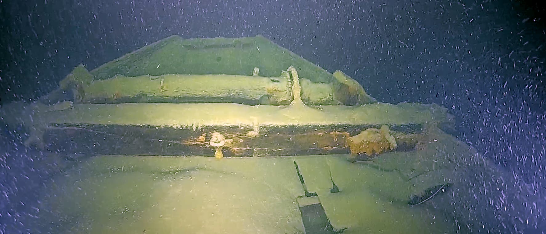 Unterwasseraufnahme eines alten Schiffswracks, das von Sand und Meeresbewuchs bedeckt ist. Die Struktur des Wracks ist teilweise sichtbar, mit erkennbaren Holzbalken und metallischen Elementen. Das Bild zeigt eine ruhige, dunkle Umgebung mit schwebenden Partikeln im Wasser, die die Tiefe und Abgeschiedenheit des Fundorts betonen.