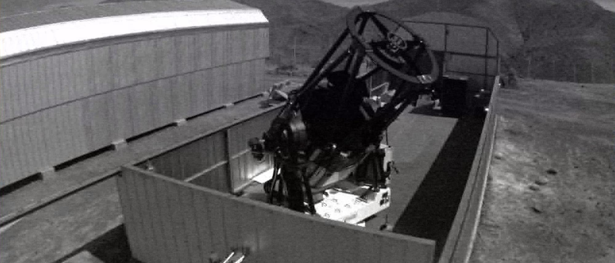 Ein gro&szlig;es Teleskop ist in einer offenen, rechteckigen Beobachtungsstation auf einem H&uuml;gel platziert. Im Hintergrund sind Berge unter einem bew&ouml;lkten Himmel zu sehen. Die Struktur des Teleskops ist komplex, mit einem gro&szlig;en, runden Spiegel und mehreren mechanischen Komponenten. Die Umgebung wirkt abgelegen und ruhig, was auf eine astronomische Beobachtungsst&auml;tte hinweist.