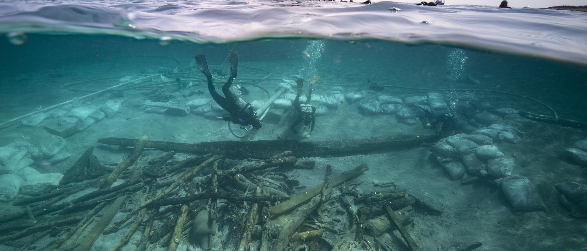 Taucher erkunden eine Unterwasser-Ausgrabungsst&auml;tte mit Holztr&uuml;mmern, Amphoren und Sands&auml;cken auf dem Meeresboden. &Uuml;ber der Wasseroberfl&auml;che ist ein Boot mit mehreren Personen zu sehen. Die Szene zeigt eine arch&auml;ologische Untersuchung in einem klaren blauen Meer.