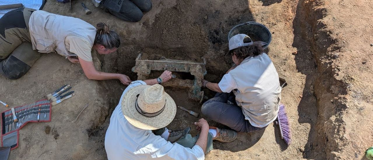 Eine Gruppe von Arch&auml;ologen arbeitet an einer Ausgrabungsst&auml;tte. Vier Personen sind in einem Graben besch&auml;ftigt, ein antikes Artefakt freizulegen. Sie tragen Schutzkleidung und H&uuml;te, um sich vor der Sonne zu sch&uuml;tzen. Werkzeuge und Ausr&uuml;stungsgegenst&auml;nde liegen um sie herum. Die Szene zeigt konzentrierte Teamarbeit in einem arch&auml;ologischen Kontext.