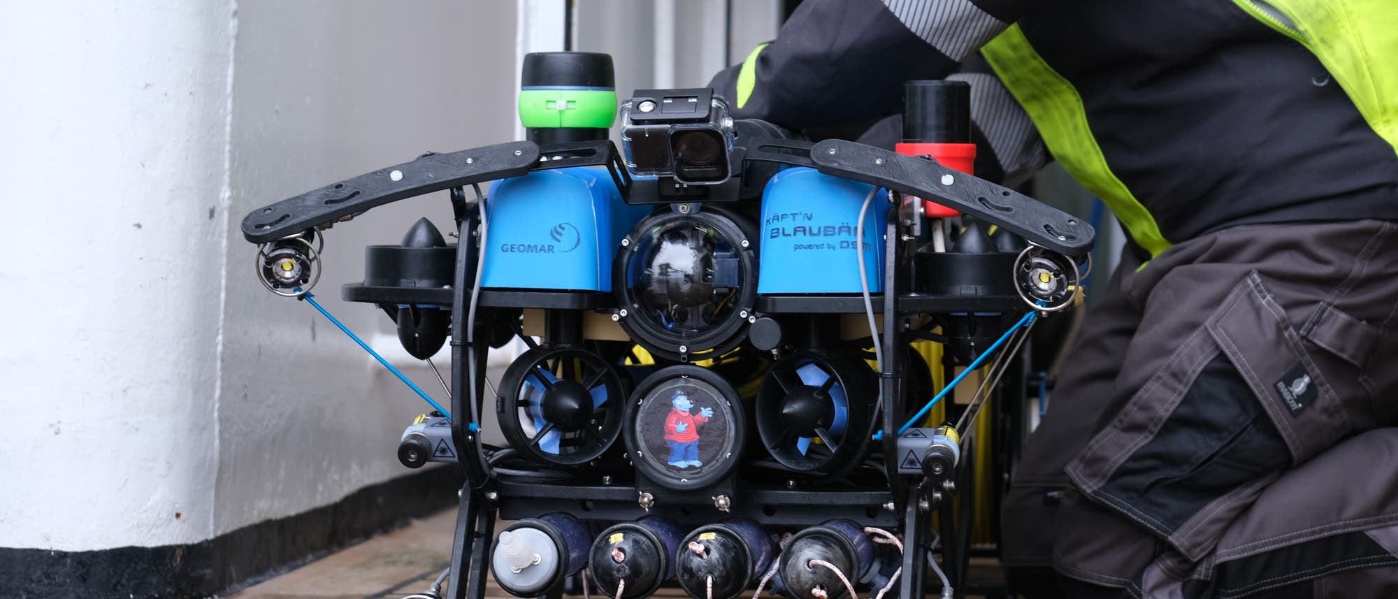 Ein Unterwasserroboter mit blauen und schwarzen Komponenten wird von einer Person in gelber Sicherheitskleidung auf einem Holzdeck vorbereitet. Der Roboter trägt die Aufschrift „GEOMAR“ und „Käpt`n Blaubär“. Er ist mit mehreren Kameras und Sensoren ausgestattet. Die Szene findet in der Nähe einer Tür statt, die zu einem Innenraum führt.