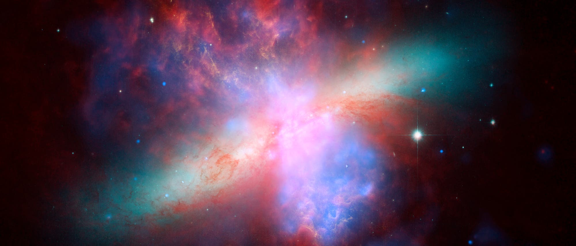 Ein farbenfrohes astronomisches Bild zeigt eine Galaxie mit leuchtenden Gaswolken in Rot, Blau und Violett. Im Zentrum befindet sich ein heller, strahlender Bereich, umgeben von diffusen Nebeln. Sterne sind im Hintergrund verstreut sichtbar. Das Bild vermittelt die Weite und Komplexit&auml;t des Universums.