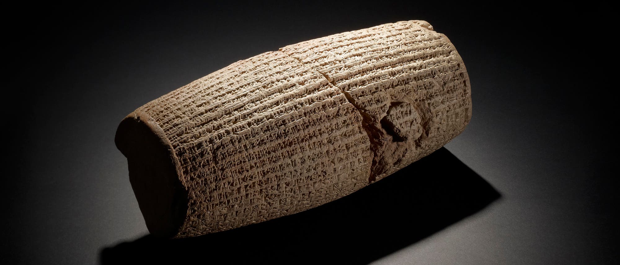 Eine antike, zylindrische Tonrolle mit eingravierten Keilschriftzeichen, die auf einem dunklen Hintergrund liegt. Die Rolle ist leicht beschädigt, was auf ihr Alter hinweist. Die Beleuchtung hebt die Textur und die Details der Inschriften hervor.