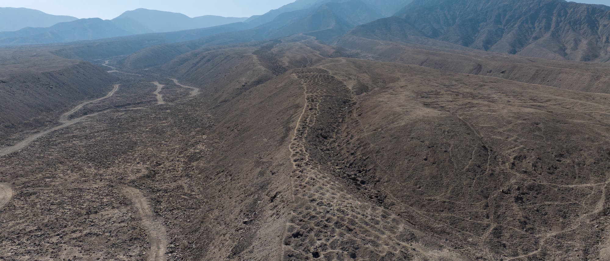 Luftaufnahme einer trockenen, hügeligen Landschaft mit erodierten Bergrücken und Tälern. Im Hintergrund sind grüne landwirtschaftliche Felder und ein Flussbett zu sehen, die einen Kontrast zur kargen Wüstenlandschaft bilden. Die Szene zeigt eine lange Reihe künstlicher Gruben.
