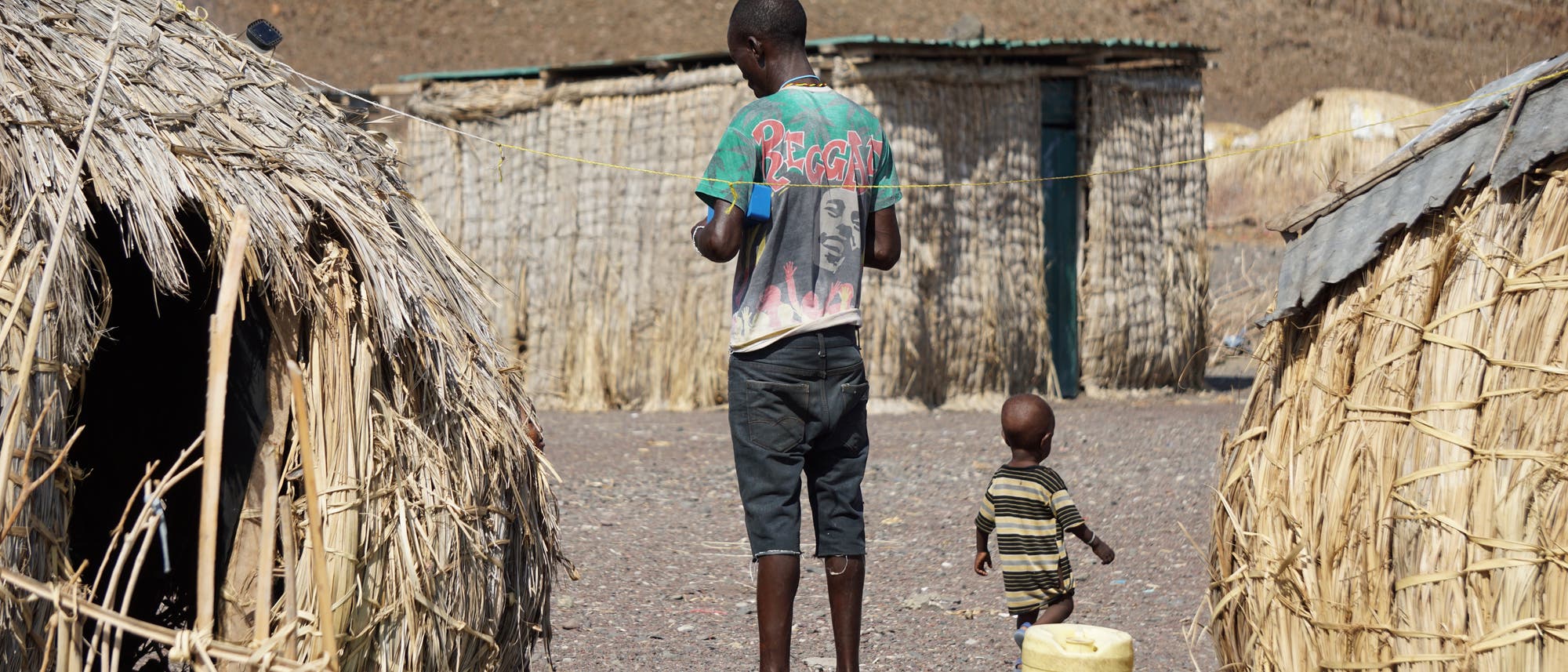 Ein Mann und ein Junge stehen inmitten von Strohhütten auf steinigem Boden.