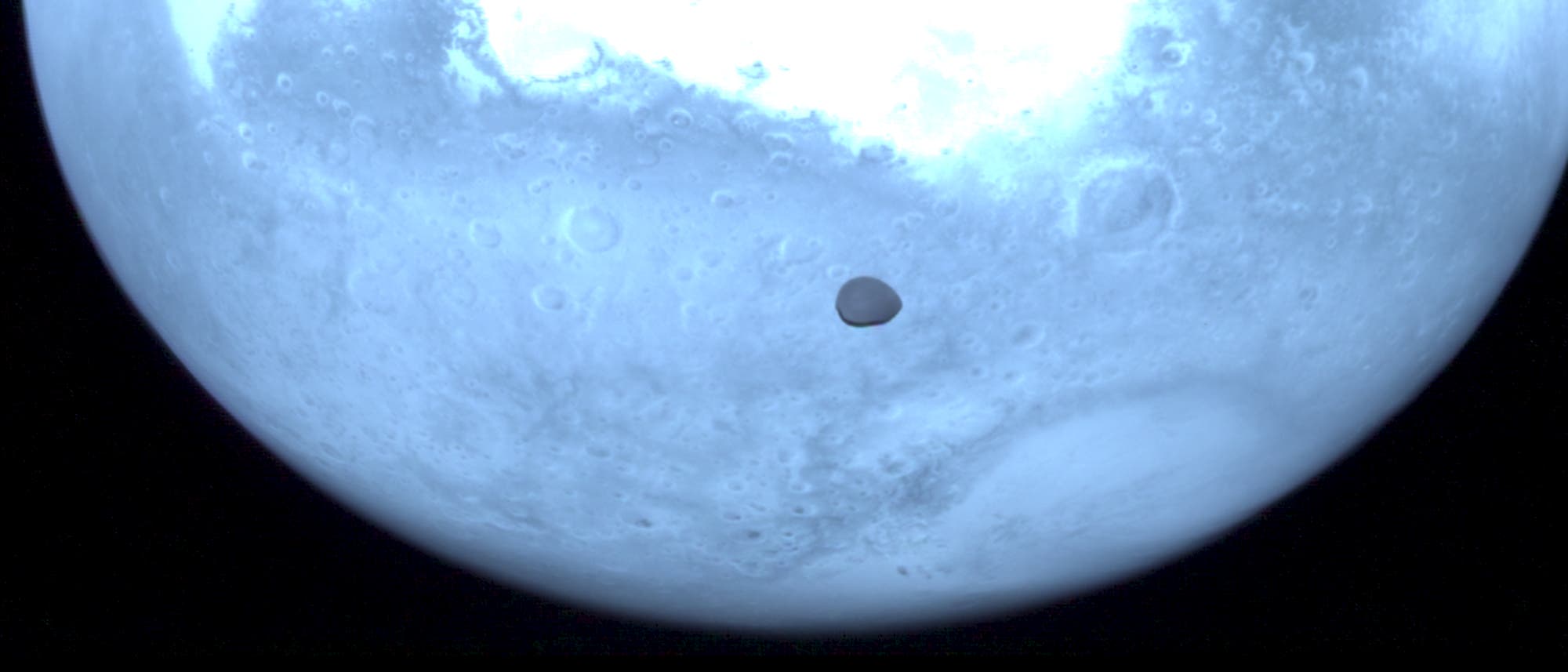 Eine Nahaufnahme des Mars mit seinem Mond Deimos, der als dunkler Schatten vor der hellen, kraterübersäten Oberfläche des Planeten vorbeizieht. Die Aufnahme zeigt die detaillierte Struktur der Marsoberfläche und die Bewegung des Mondes. Eine Nahaufnahme des Mars mit seinem Mond Deimos, der als dunkler Schatten vor der hellen, kraterübersäten Oberfläche des Planeten vorbeizieht. Die Aufnahme zeigt die detaillierte Struktur der Marsoberfläche und die Bewegung des Mondes.