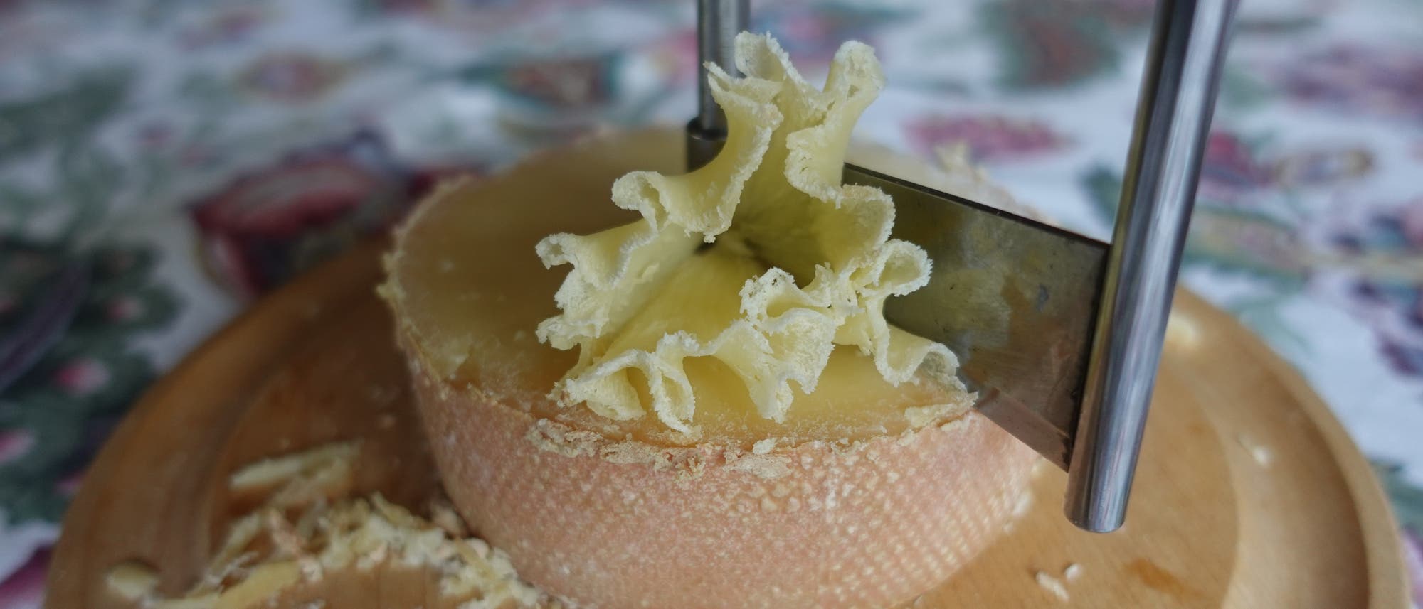 Ein runder Laib Tête-de-Moine-Käse wird auf einem Holzbrett mit einer Girolle geschnitten. Die Girolle erzeugt feine, blumenartige Käserosetten. Im Hintergrund ist ein gemustertes Tischtuch zu sehen.