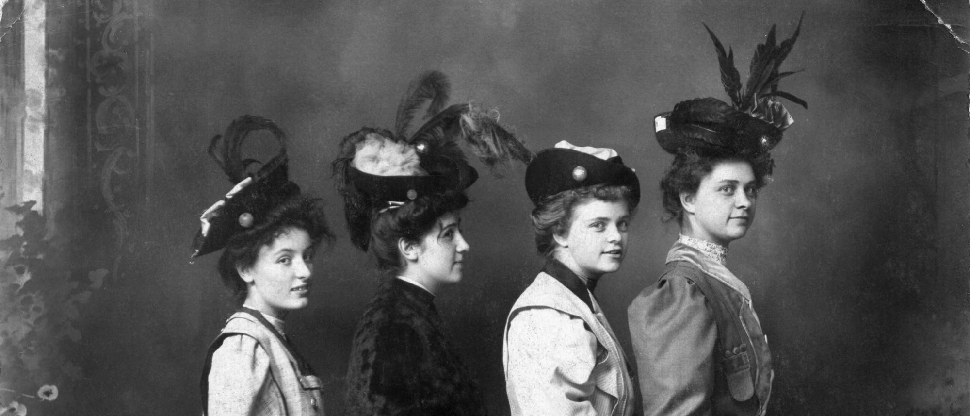 Eine historische Schwarz-Wei&szlig;-Fotografie zeigt vier Frauen in einer Reihe,. Sie tragen elegante Kleidung und H&uuml;te mit Federn, typisch f&uuml;r die Mode des fr&uuml;hen 20. Jahrhunderts. Der Hintergrund ist neutral, was den Fokus auf die Personen lenkt. Die Frauen blicken in dieselbe Richtung, was eine harmonische und stilvolle Komposition erzeugt.
