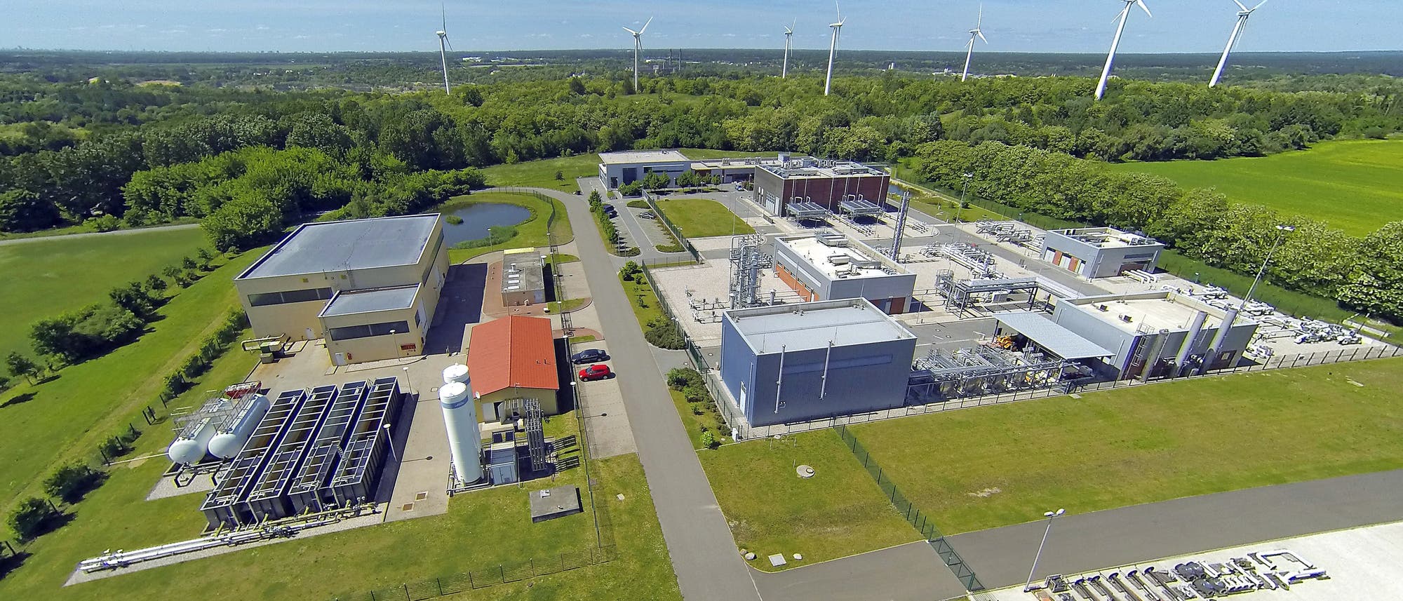 Luftaufnahme einer industriellen Anlage, umgeben von grünen Feldern und Bäumen. Im Hintergrund sind mehrere Windkraftanlagen zu sehen. Die Anlage besteht aus mehreren Gebäuden und technischen Strukturen, die durch Straßen verbunden sind. Ein kleiner Teich befindet sich in der Nähe der Gebäude. Der Himmel ist klar und blau.