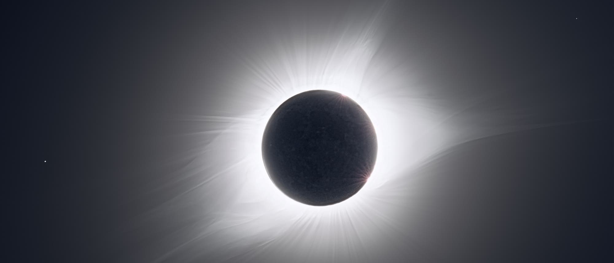 Eine totale Sonnenfinsternis, bei der der Mond die Sonne vollst&auml;ndig verdeckt. Die Sonnenkorona ist als leuchtender Ring um den Mond sichtbar, mit strahlenf&ouml;rmigen Lichtmustern, die sich in den dunklen Himmel erstrecken. Links im Bild ist ein kleiner Lichtpunkt zu sehen, m&ouml;glicherweise ein Stern oder Planet.