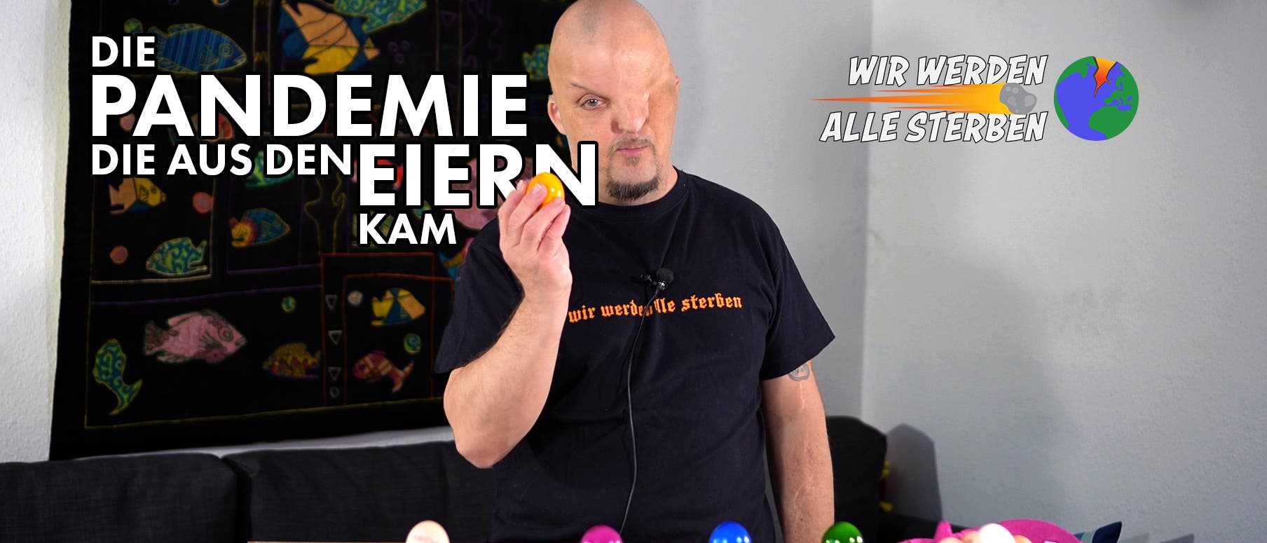 Eine Person steht in einem Raum und hält ein gelbes Ei in der Hand. Auf der linken Seite des Bildes steht der Text: "Die Pandemie, die aus den Eiern kam". Auf der rechten Seite ist ein Logo mit der Aufschrift "Wir werden alle sterben" und einer stilisierten Weltkugel zu sehen. Auf dem Tisch vor der Person stehen mehrere bunte Eier in Eierbechern. Im Hintergrund hängt ein Wandteppich mit bunten Fischen. Die Person trägt ein schwarzes T-Shirt mit dem Text "wir werden alle sterben".