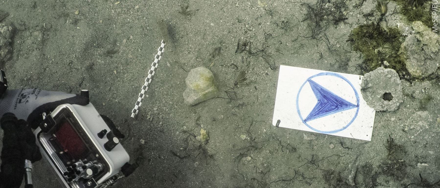 Unterwasseraufnahme eines sandigen Meeresbodens mit einem St&uuml;ck Marmor und einer Kamera im Vordergrund. Auf dem Boden liegt eine wei&szlig;e Tafel mit einem blauen Kreis und einem darin gezeichneten blauen Pfeil. Daneben befindet sich ein Ma&szlig;stab. Um die Tafel herum sind Algen und Steine zu sehen.