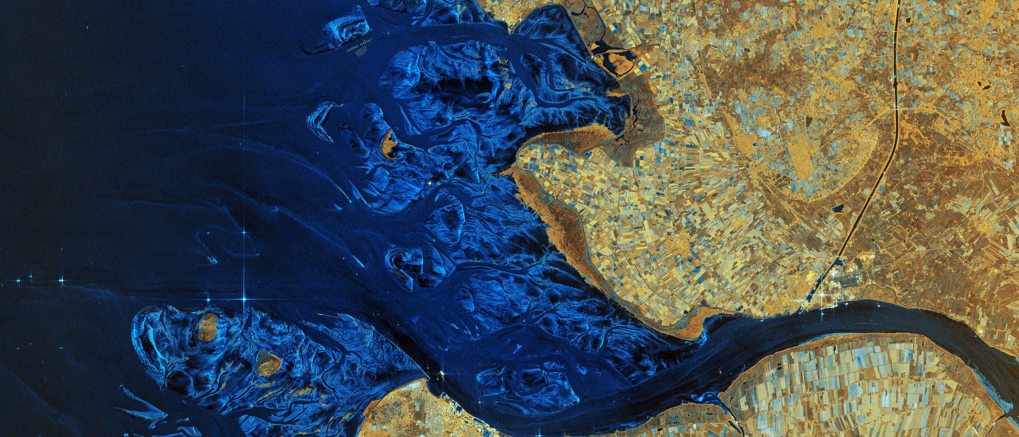 Satellitenbild einer Küstenregion mit dem Flussdelta der Elbe, die in ein tiefblaues Meer mündet. Die linke Bildhälfte zeigt das Meer mit verzweigten Wasserwegen, während die rechte Bildhälfte landwirtschaftlich genutzte Flächen in verschiedenen Brauntönen darstellt. Die Karte hebt die Kontraste zwischen Wasser und Land hervor und zeigt die natürliche Geografie der Region.