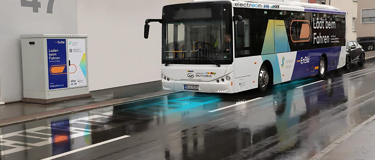 Ein Elektrobus fährt auf einer nassen Straße vor einem Gebäude mit der Nummer 47. Der Bus ist mit der Aufschrift "Laden beim Fahren" versehen und nutzt eine induktive Ladetechnologie, die durch blaue Markierungen auf der Straße angezeigt wird. Links im Bild befindet sich ein Schaltkasten mit der Aufschrift "Laden beim Fahren" und einem QR-Code. Der Bus gehört zum Projekt "e-GELU" und ist mit Logos verschiedener Partner versehen. Im Hintergrund sind moderne Wohngebäude zu sehen. Ein Elektrobus fährt auf einer nassen Straße vor einem Gebäude mit der Nummer 47. Der Bus ist mit der Aufschrift "Laden beim Fahren" versehen und nutzt eine induktive Ladetechnologie, die durch blaue Markierungen auf der Straße angezeigt wird. Links im Bild befindet sich ein Schaltkasten mit der Aufschrift "Laden beim Fahren" und einem QR-Code. Der Bus gehört zum Projekt "e-GELU" und ist mit Logos verschiedener Partner versehen. Im Hintergrund sind moderne Wohngebäude zu sehen.