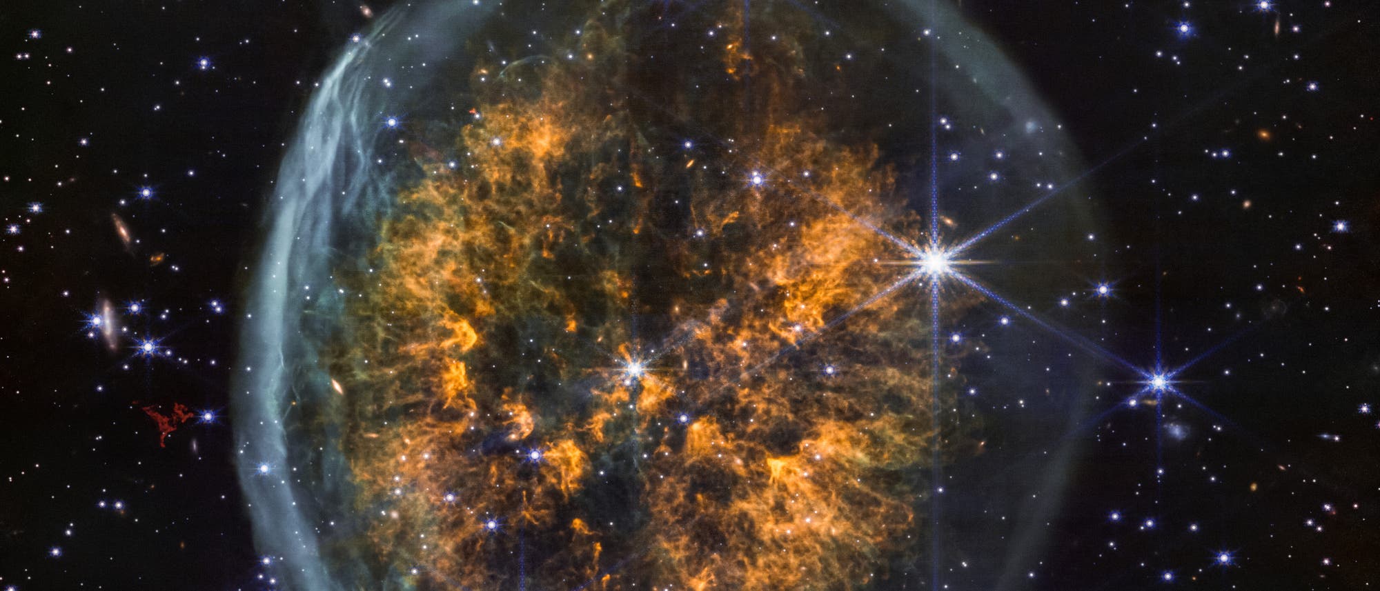 Ein astronomisches Bild zeigt einen planetarischen Nebel im Weltraum. Der Nebel hat eine blasenartige Form mit einem leuchtenden, orangefarbenen Zentrum, das von einem bl&auml;ulichen, durchscheinenden Rand umgeben ist. Im Hintergrund sind zahlreiche Sterne und Galaxien zu sehen, die als helle Punkte und spiralf&ouml;rmige Strukturen erscheinen. Das Bild vermittelt die Weite und Komplexit&auml;t des Universums.