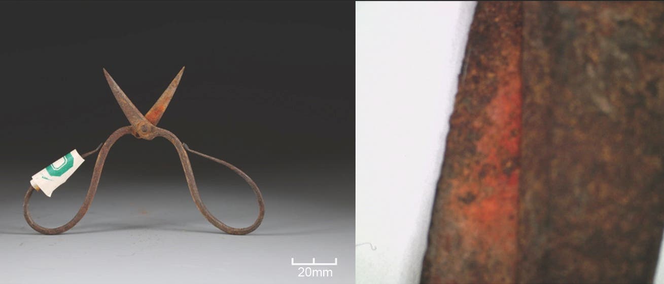 Eine rostige, altertümliche Schere mit gebogenen Griffen und spitzen Klingen ist auf der linken Seite des Bildes zu sehen. Ein kleines Etikett mit einem grünen Symbol ist an einem Griff befestigt. Rechts im Bild befindet sich eine Nahaufnahme der Oberfläche der Schere, die eine rötliche Verfärbung zeigt. Unten links ist eine Maßstabsleiste von 20 mm abgebildet. Der Hintergrund ist dunkel, um die Details der Schere hervorzuheben. Eine rostige, altertümliche Schere mit gebogenen Griffen und spitzen Klingen ist auf der linken Seite des Bildes zu sehen. Ein kleines Etikett mit einem grünen Symbol ist an einem Griff befestigt. Rechts im Bild befindet sich eine Nahaufnahme der Oberfläche der Schere, die eine rötliche Verfärbung zeigt. Unten links ist eine Maßstabsleiste von 20 mm abgebildet. Der Hintergrund ist dunkel, um die Details der Schere hervorzuheben.