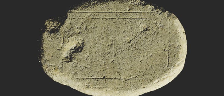 Ein ovaler, flacher Stein mit einer rauen, sandfarbenen Oberfl&auml;che. Auf dem Stein sind feine, rechteckige Linien eingraviert, die m&ouml;glicherweise eine alte Inschrift oder Markierung darstellen. Der Hintergrund ist dunkel, wodurch der Stein hervorgehoben wird.