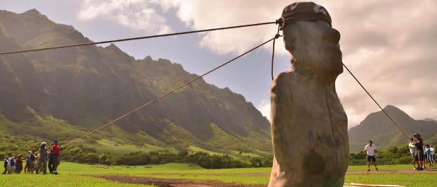 Eine Gruppe von Menschen zieht an Seilen, die an einer großen Statue befestigt sind, um sie aufrecht zu halten. Die Szene spielt sich auf einer grünen Wiese vor einer beeindruckenden Bergkulisse ab. Der Himmel ist teilweise bewölkt, und die Sonne scheint. Die Menschen scheinen an einer Demonstration oder einem Experiment teilzunehmen, das möglicherweise die Bewegung oder Stabilität der Statue untersucht.