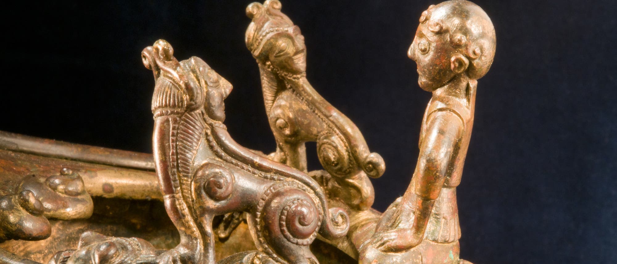 Eine kunstvolle Bronzeskulptur zeigt eine Figur, die auf einem aufwendig verzierten Objekt steht. Die Skulptur enthält detaillierte, spiralförmige Muster und stilisierte Tierköpfe. Die Figur scheint in Interaktion mit den Tierköpfen zu stehen, was auf eine symbolische oder mythologische Bedeutung hindeutet. Die Oberfläche der Skulptur weist eine antike Patina auf, die auf ihr Alter hinweist. Eine kunstvolle Bronzeskulptur zeigt eine Figur, die auf einem aufwendig verzierten Objekt steht. Die Skulptur enthält detaillierte, spiralförmige Muster und stilisierte Tierköpfe. Die Figur scheint in Interaktion mit den Tierköpfen zu stehen, was auf eine symbolische oder mythologische Bedeutung hindeutet. Die Oberfläche der Skulptur weist eine antike Patina auf, die auf ihr Alter hinweist.