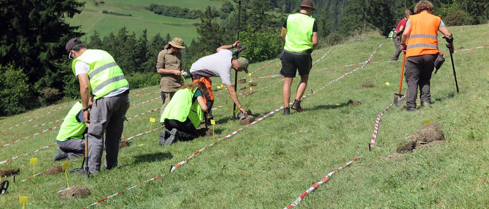 Eine Gruppe von Personen in Warnwesten arbeitet auf einer Wiese, die mit rot-weißem Absperrband markiert ist. Einige knien und graben mit kleinen Werkzeugen im Boden, während andere stehen und Werkzeuge halten. Die Umgebung ist ländlich mit Hügeln und Bäumen im Hintergrund. Die Szene deutet auf eine archäologische Untersuchung hin. Eine Gruppe von Personen in Warnwesten arbeitet auf einer Wiese, die mit rot-weißem Absperrband markiert ist. Einige knien und graben mit kleinen Werkzeugen im Boden, während andere stehen und Werkzeuge halten. Die Umgebung ist ländlich mit Hügeln und Bäumen im Hintergrund. Die Szene deutet auf eine archäologische Untersuchung hin.