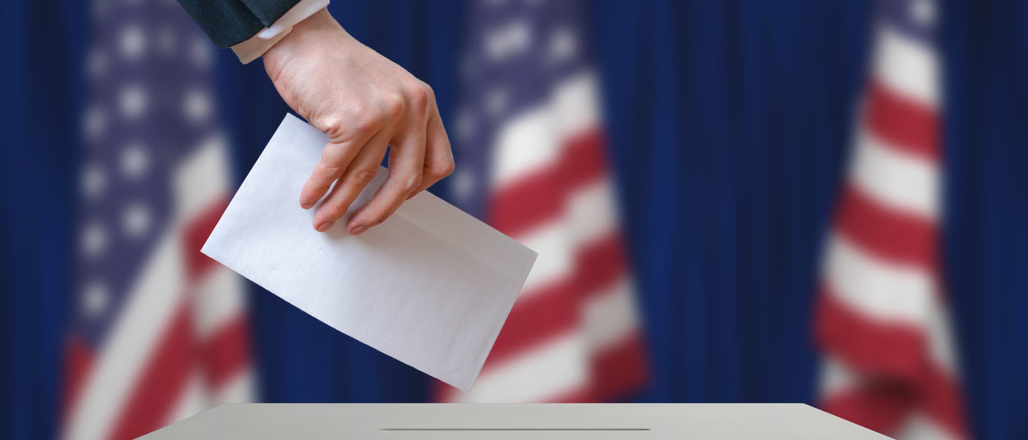 Eine Hand wirft einen Zettel in eine Wahlurne. Im Hintergrund drei US-Flaggen