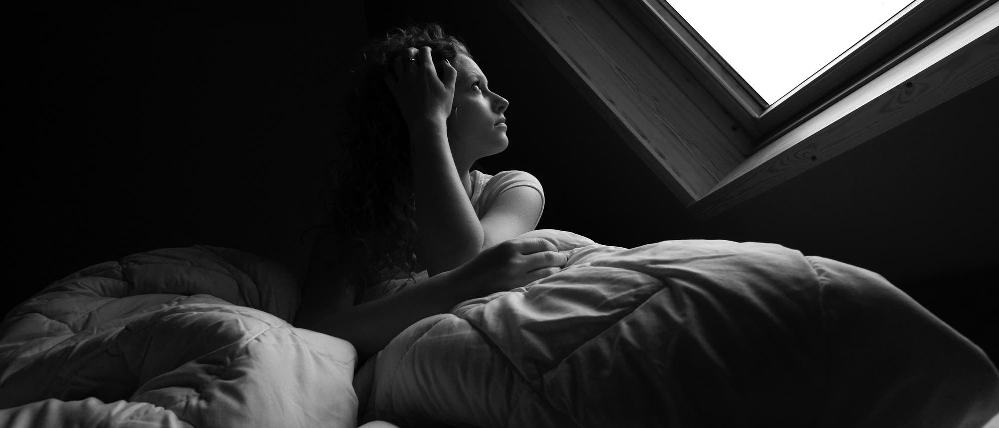 Eine Frau liegt im Bett und sieht durch ein Dachfenster in den grauen Himmel.