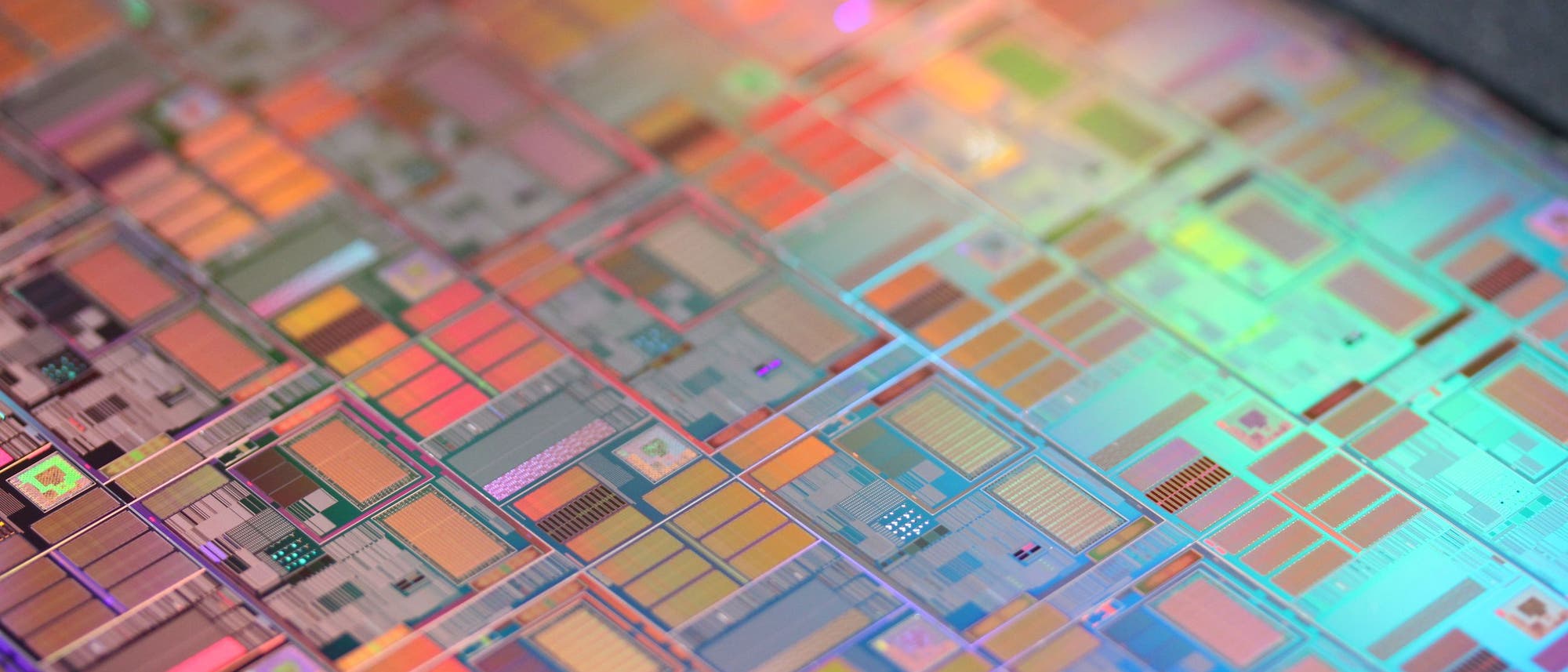 Eine Nahaufnahme eines Silizium-Wafers mit zahlreichen Mikrochip-Schaltkreisen. Die Oberfl&auml;che zeigt ein komplexes Muster aus rechteckigen und quadratischen Formen in verschiedenen Farben, die durch die Reflexion des Lichts entstehen. Der Wafer ist leicht geneigt, was einen Teil des Hintergrunds sichtbar macht.
