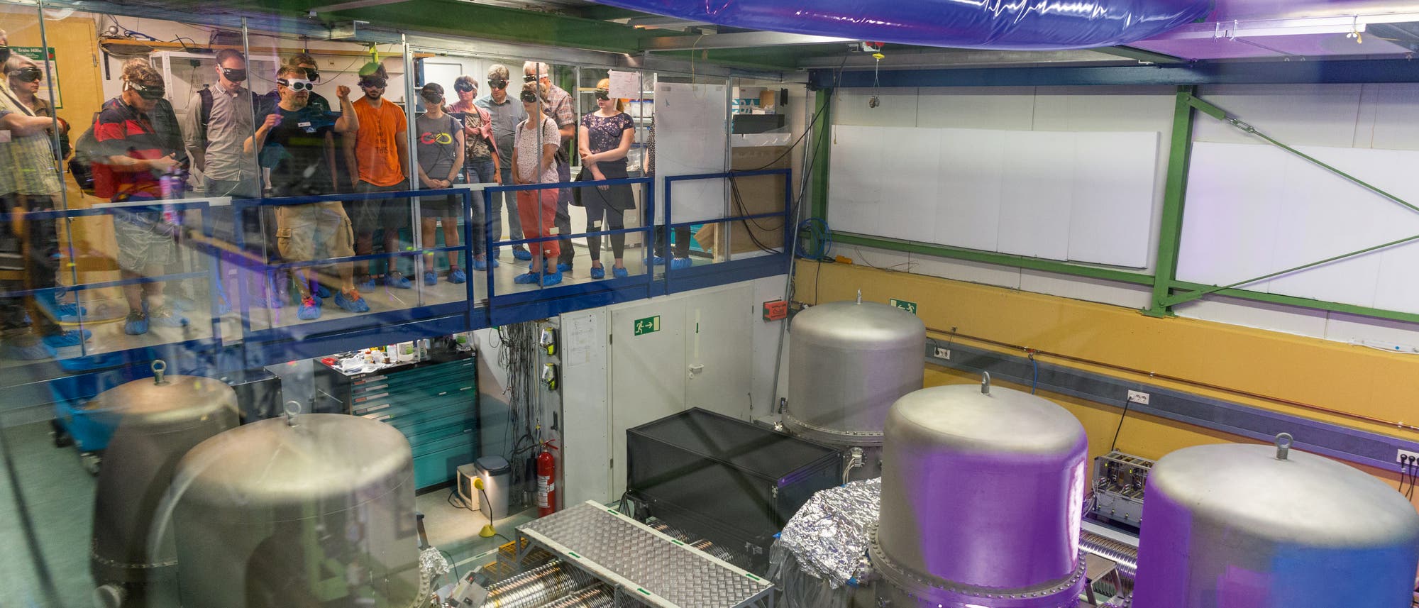 Eine Gruppe von Menschen steht auf einer erhöhten Plattform und beobachtet eine industrielle Anlage mit großen, zylinderförmigen Maschinen und technischen Geräten. Die Szene befindet sich in einem Labor oder einer Fabrikhalle mit metallischen Strukturen und Rohren. Die Personen tragen Schutzbrillen und Helme, was auf eine Sicherheitsvorschrift hinweist. Im Vordergrund sind mehrere große, silberne Tanks und komplexe Maschinen zu sehen. Die Umgebung wirkt technisch und industriell. Eine Gruppe von Menschen steht auf einer erhöhten Plattform und beobachtet eine industrielle Anlage mit großen, zylinderförmigen Maschinen und technischen Geräten. Die Szene befindet sich in einem Labor oder einer Fabrikhalle mit metallischen Strukturen und Rohren. Die Personen tragen Schutzbrillen und Helme, was auf eine Sicherheitsvorschrift hinweist. Im Vordergrund sind mehrere große, silberne Tanks und komplexe Maschinen zu sehen. Die Umgebung wirkt technisch und industriell.